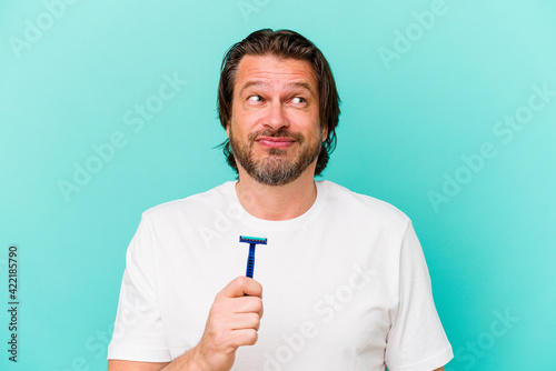 Middle age dutch man holding a razor blade isolated on blue background confused, feels doubtful and unsure.