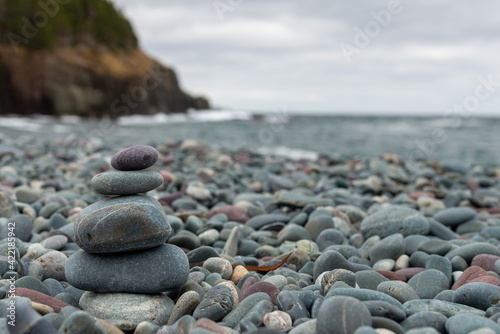 A small rock inukshuk or stack on a wide open beach. The Inuit rock symbolizes direction. The beach has a landmass in the background with trees and rock cliffs. The sky is grey with thick clouds.