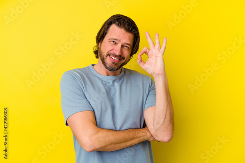 Middle age caucasian man isolated on yellow background winks an eye and holds an okay gesture with hand.