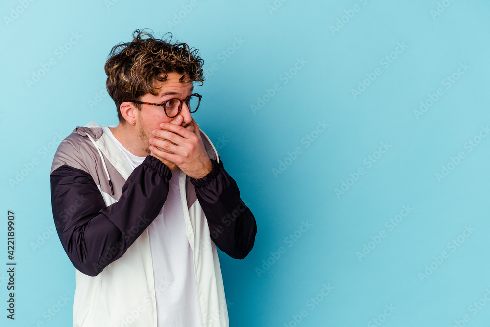 Young caucasian man wearing eyeglasses isolated on blue background thoughtful looking to a copy space covering mouth with hand. Young caucasian man wearing eyeglasses isolated on blue background thoughtful looking to a copy space covering mouth with hand.