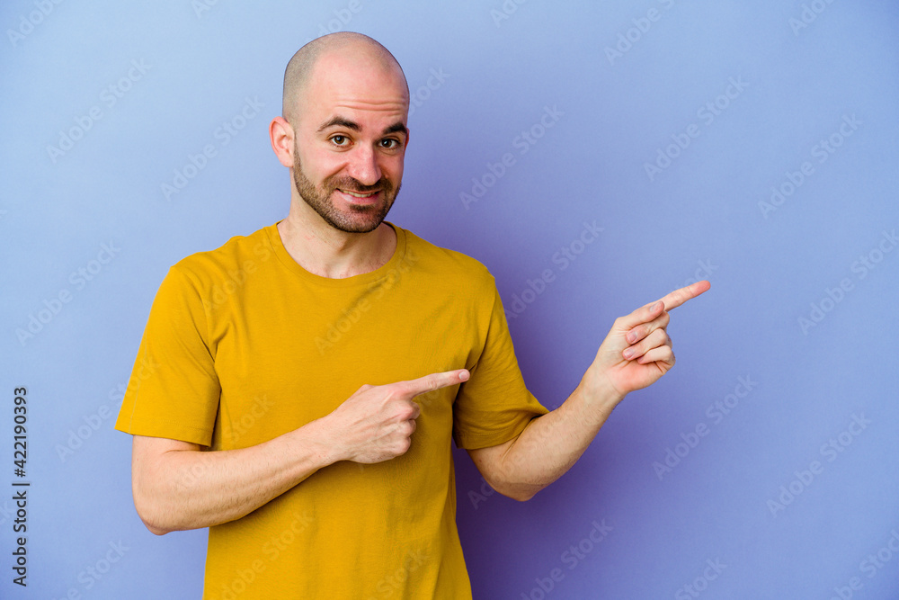 Young caucasian bald man isolated on purple background shocked pointing with index fingers to a copy space. Young caucasian bald man isolated on purple background shocked pointing with index fingers to a copy space.