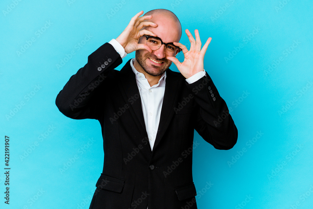 Young business caucasian bald man isolated on blue background showing okay sign over eyes Young business caucasian bald man isolated on blue background showing okay sign over eyes