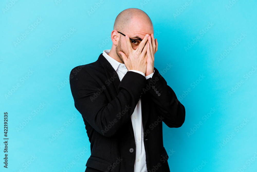 Young business caucasian bald man isolated on blue background blink through fingers frightened and nervous. Young business caucasian bald man isolated on blue background blink through fingers frightened and nervous.