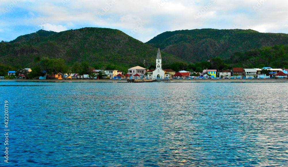 Caribbean village on a quiet cloudy day: town dock, waterfront and church with a bell and a spire, small local colorful homes; view from the deck of a charter boat.