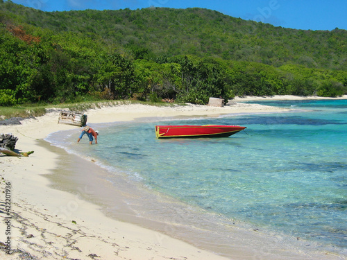 Tropical beach in St Vincent & the Grenadines, white coral sand, blue ocean water and lush green tropical jungle, a red boat on the water, a tourist collecting shells.