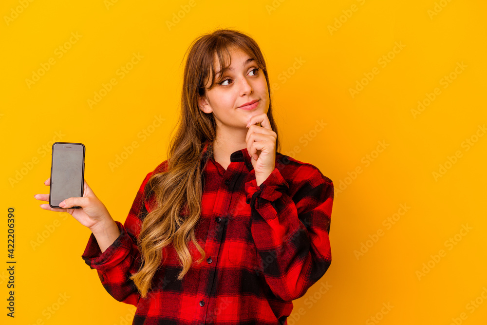 Young caucasian woman holding phone isolated on yellow background looking sideways with doubtful and skeptical expression. Young caucasian woman holding phone isolated on yellow background looking sideways with doubtful and skeptical expression.