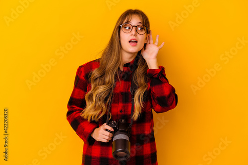 Young photographer caucasian woman isolated on yellow background trying to listening a gossip.
