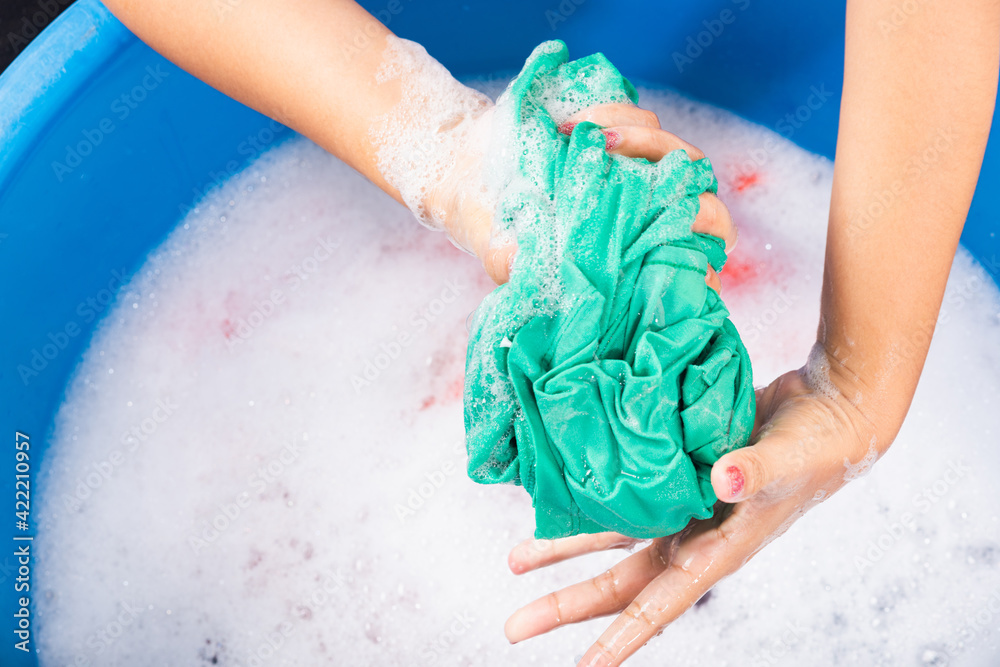 Fototapeta premium Closeup young Asian woman use hands washing color clothes in basin. Female squeeze wring out wet fabric cloth with detergent have soapy bubble in water, studio shot background, laundry concept