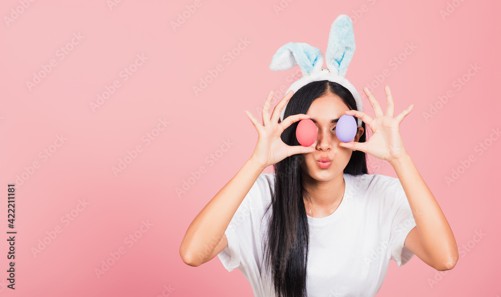 Happy beautiful young woman smiling wearing rabbit ears holding colorful Easter eggs front eyes, Thai female with bunny ear hold easter egg covering eye, studio shot isolated on pink background