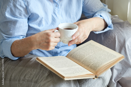Close-up of a mature woman reading a book at home by the window, sitting on the sofa with a cup of coffee.