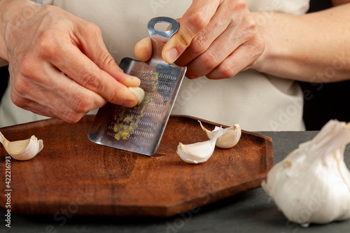 Quadro em tela Hands of a caucasian woman grating garlic cloves using shovel shaped metal mini grater on a wooden plate
