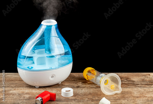 asthma, allergy airway problem, pulmonary disorders concept with ultrasonic tabletop humidifier creating steam. Inhaler and chamber mask to deliver emergency medication are left on tabletop. photo