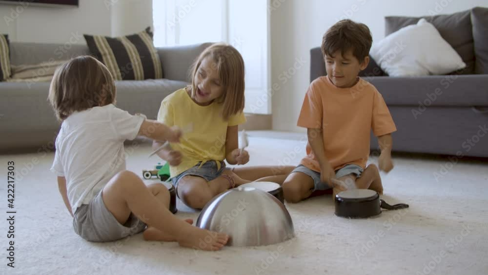 Happy little kids playing in living room with utensils. Blonde girl and two boys smiling, sitting on floor together and knocking on pans. Static camera. Childhood, noise and home activity concept