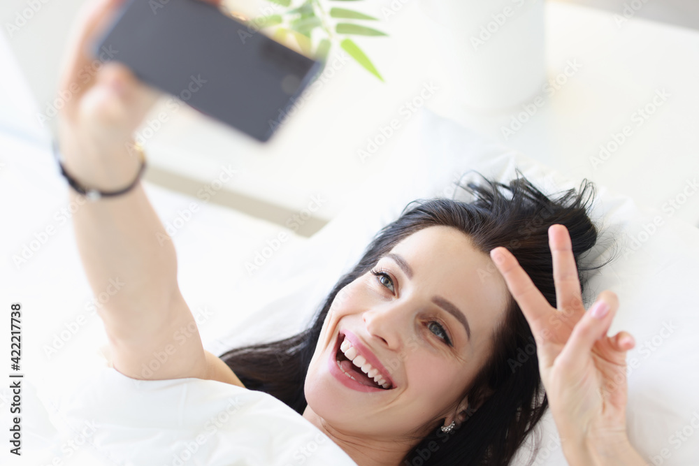 Fototapeta premium Portrait of smiling young woman on white bed taking selfie on smartphone