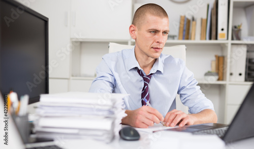 Portrait of manager dressed in the shirt with a tie working with documents and laptop at the modern office interior