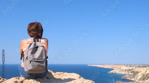 Young happy woman with a backpack sits on a rock and looks at the valley below. Tourists on the rocky coast of the Black Sea. The concept of freedom and ease