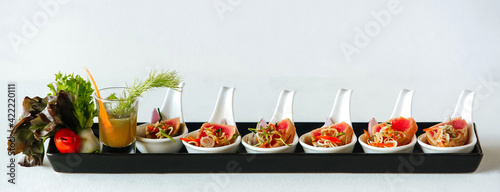 Snacks in white spoons with red potatoes or meat, with vegetables and sauce in a shot glass on long plate on white background 