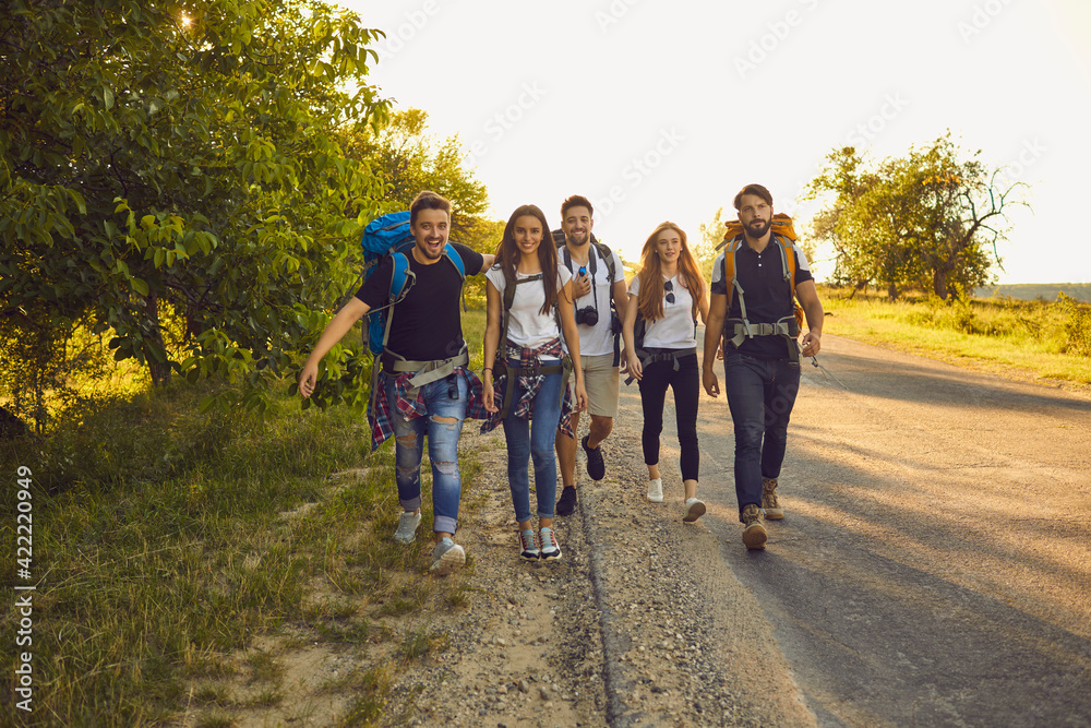 Group of happy young friends with backpacks walking along road in countryside. Hikers on tourist trip in summertime Group of happy young friends with backpacks walking along road in countryside. Hikers on tourist trip in summertime