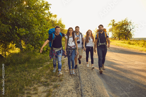 Group of happy young friends with backpacks walking along road in countryside. Hikers on tourist trip in summertime