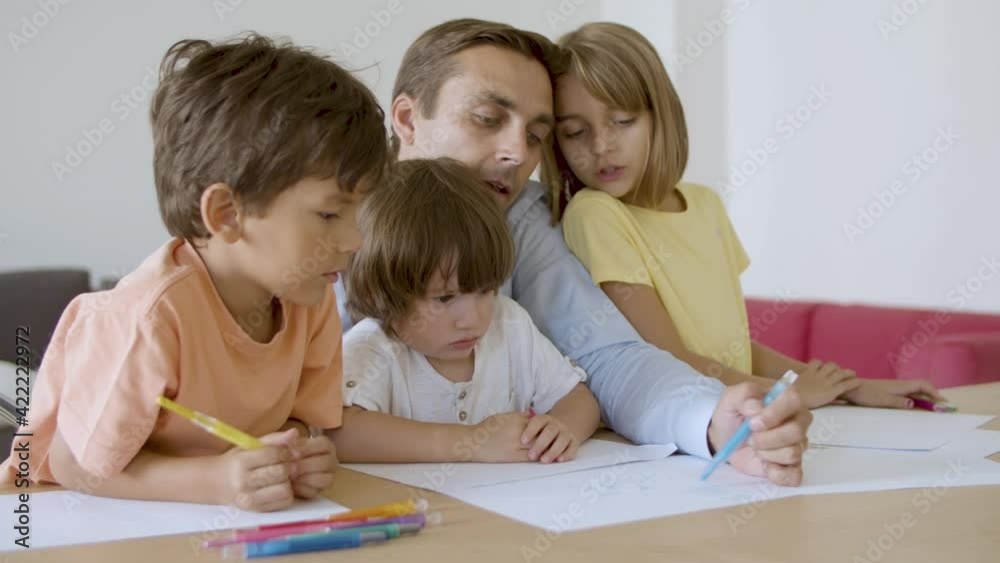 Caring dad and lovely kids drawing with marker on paper. Concentrated Caucasian father painting with pen and playing with children in living room. Static camera. Fatherhood and family time concept Caring dad and lovely kids drawing with marker on paper. Concentrated Caucasian father painting with pen and playing with children in living room. Static camera. Fatherhood and family time concept