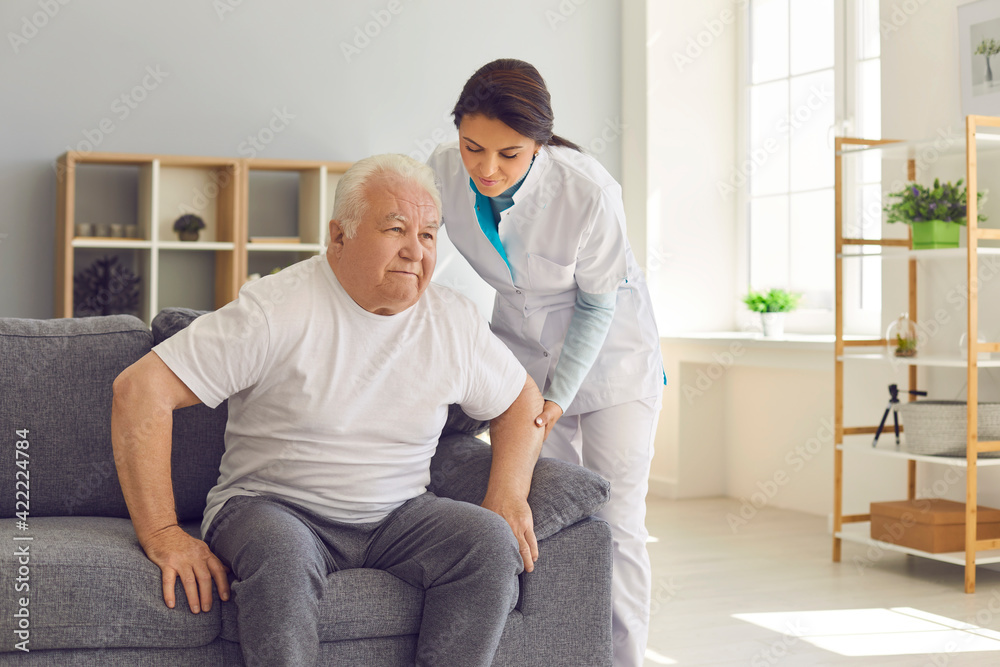 Friendly female doctor helps an older man who is trying to get up from the couch. Friendly female doctor helps an older man who is trying to get up from the couch.