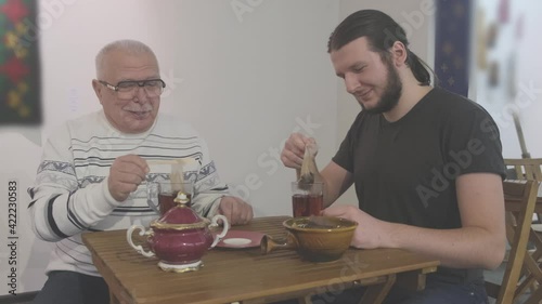 Young man with beard and sideburns in black T-shirt and pensioner in glasses and warm sweater talk while making tea in cafe