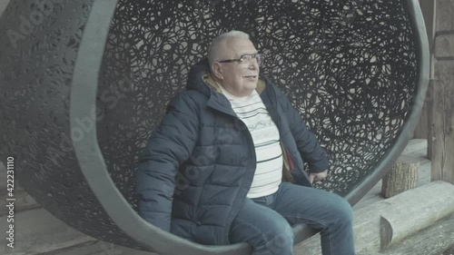 Senior gray haired man in winter jacket warm sweater and denim jeans sits in hanging cocoon chair against wooden house closeup