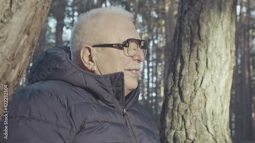 Smiling grey-haired senior man in glasses and black winter jacket looks straight against spruce and aspen on sunny day closeup