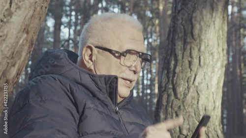 Elderly man with gray mustache in glasses and black winter jacket dials number on smartphone calling against forest closeup