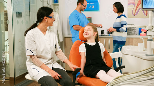 Little girl patient waiting pediatric stomatologist woman sitting in dental chair while man assistant talking with mother in background. Dentist discussing with girl before examining oral health.