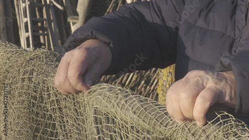 Senior man hands with wrinkles straighten stacking up old greenish fishing net hanging on rough log on sunny day closeup