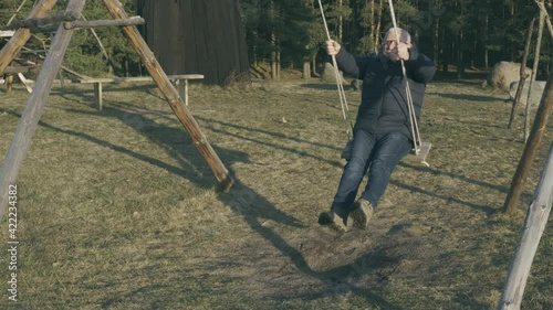 Smiling elderly man in glasses and black winter jacket rocks on swings holding ropes against forest and big stones on ground