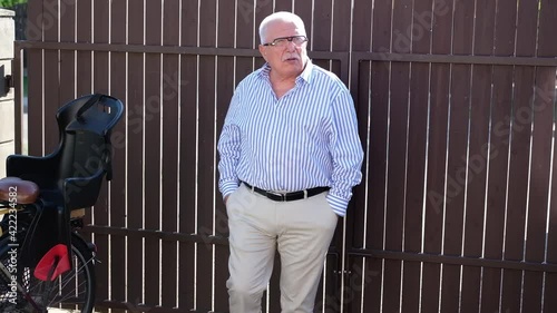 Elegant senior man in glasses in striped shirt and beige pants with black belt speaks at wooden fence and child seat on bike closeup