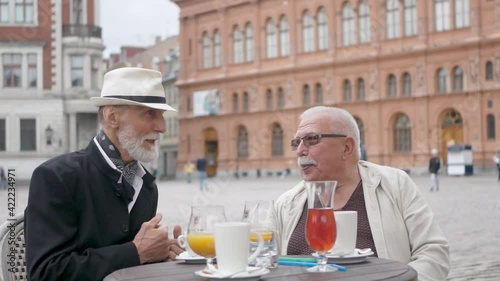 Grey haired pensioners sit in outdoor city cafe at wooden table drinking juice and talk against historic buildings and people