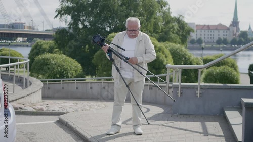 Elderly man installs camera tripod for shooting on stairs of river embankment against trees bridge buildings and old church tower