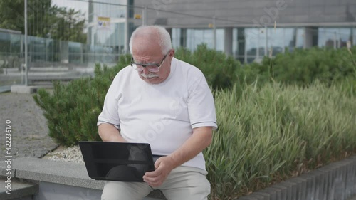 Elderly man in glasses and white trousers sits on bench holding laptop on knees and works at green bushes and building closeup