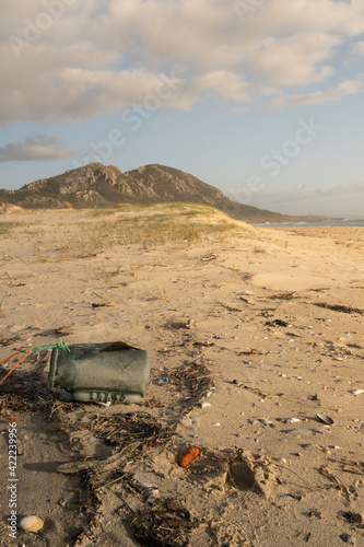 Bote de plástico (basura) en una preciosa playa con dunas y montañas al fondo, con luz de atardecer y el mar azul.