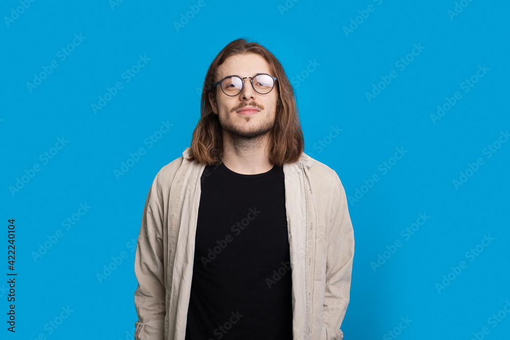 Confidend long haired man with beard is looking at camera wearing glasses and shirt on a blue studio wall Confidend long haired man with beard is looking at camera wearing glasses and shirt on a blue studio wall