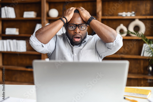 African American businessman received email with bad news, sitting at the desk with laptop, lost business, office worker made mistake, feeling scared and stressed, holding head in hands and screaming photo