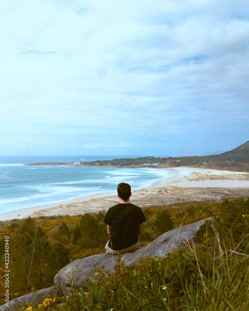 Hombre joven de espaldas sentado en una roca observando una gran playa de arena blanca con olas. 