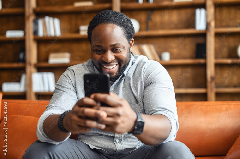 Happy laughing mixed-race man sitting on the couch, staying at home,  texting chatting with friends on mobile phone, using dating app, flirting,  watching comedy show, funny video, playing games online Stock Photo |