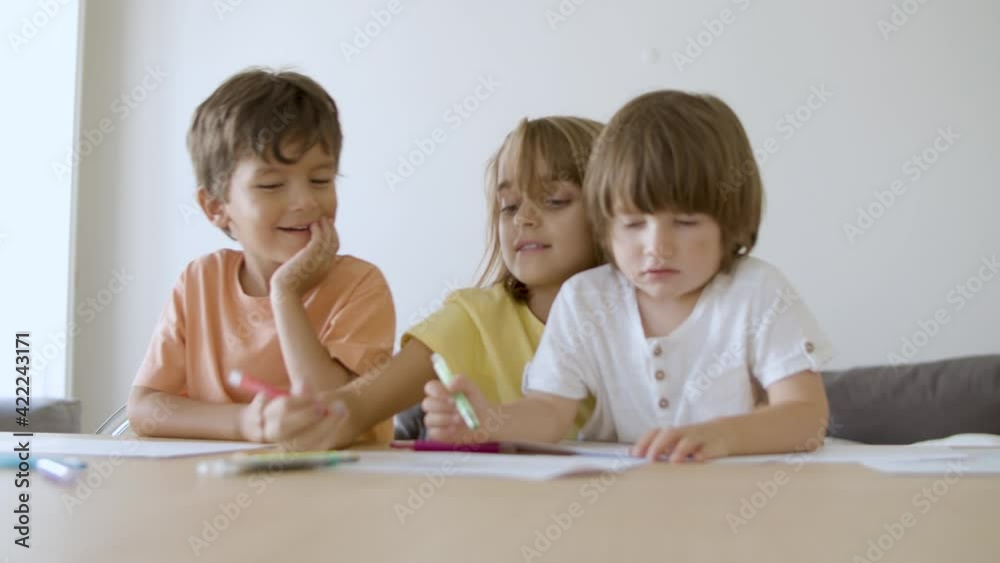 Little children painting with markers at home. Cute blonde girl holding and talking with brothers. Lovely kids sitting at table and drawing on paper. Slider motion. Childhood and creativity concept Little children painting with markers at home. Cute blonde girl holding and talking with brothers. Lovely kids sitting at table and drawing on paper. Slider motion. Childhood and creativity concept