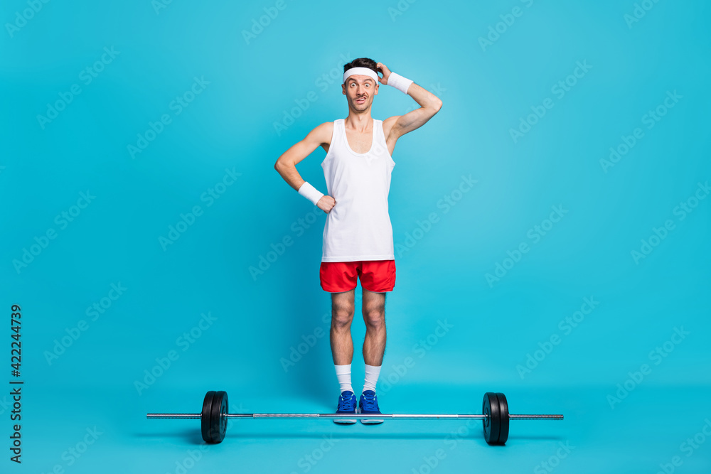 Full length photo of clueless young man scratch hand head dumbbell lift isolated on blue color background Full length photo of clueless young man scratch hand head dumbbell lift isolated on blue color background