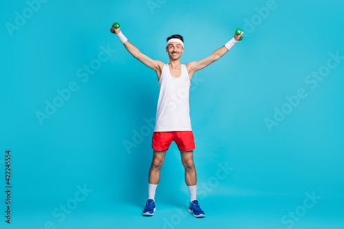 Full length photo of young sportsman happy positive smile do physical exercises with small barbells isolated over blue color background © deagreez