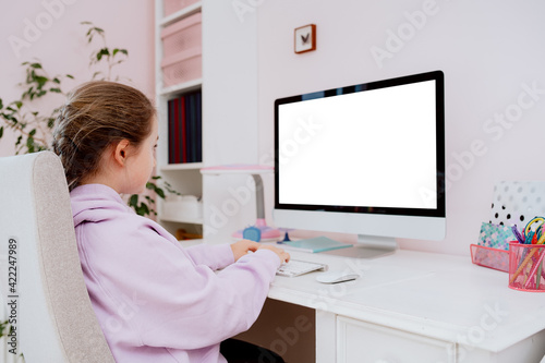 A view from behind the back of a teenage school girl who uses a desktop computer while sitting at a desk in her room. She participates in remote learning and checks content posted on social media.