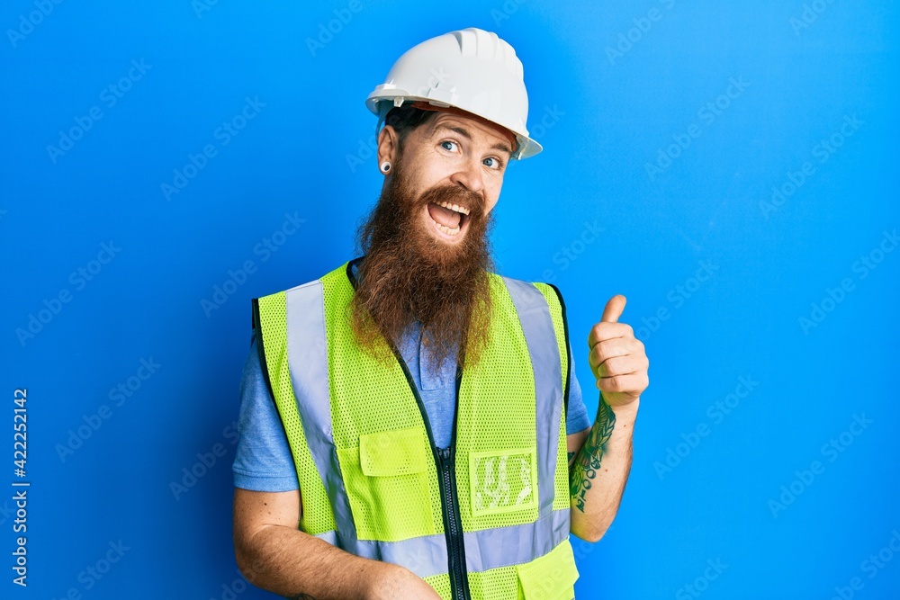 Redhead man with long beard wearing safety helmet and reflective jacket smiling with happy face looking and pointing to the side with thumb up.