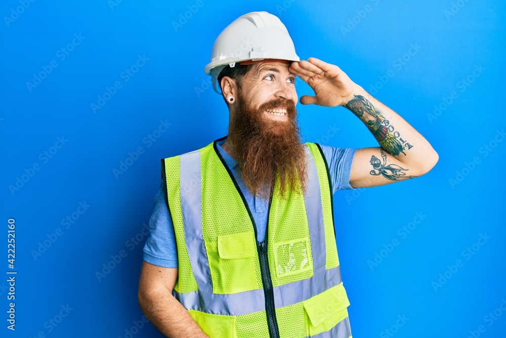 Redhead man with long beard wearing safety helmet and reflective jacket very happy and smiling looking far away with hand over head. searching concept. Redhead man with long beard wearing safety helmet and reflective jacket very happy and smiling looking far away with hand over head. searching concept.