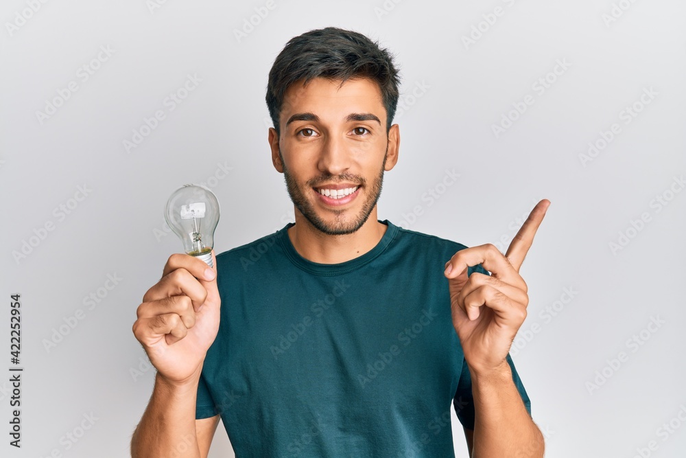 Young handsome man holding lightbulb for inspiration and idea smiling happy pointing with hand and finger to the side Young handsome man holding lightbulb for inspiration and idea smiling happy pointing with hand and finger to the side
