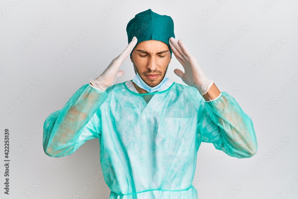 Young handsome man wearing surgeon uniform and medical mask with hand on head, headache because stress. suffering migraine. Young handsome man wearing surgeon uniform and medical mask with hand on head, headache because stress. suffering migraine.