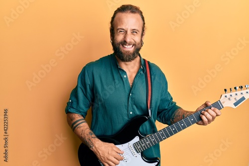 Handsome man with beard and long hair playing electric guitar smiling and laughing hard out loud because funny crazy joke.